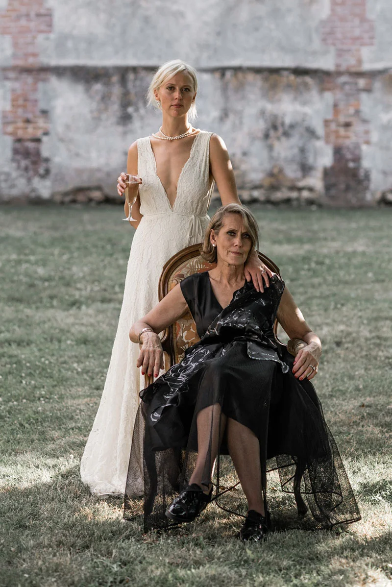 Une mariée en robe blanche et une femme en robe noire posent avec élégance devant un mur ancien.