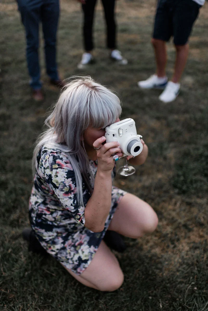 Une femme aux cheveux argentés en robe fleurie accroupie pour prendre une photo avec un appareil blanc.