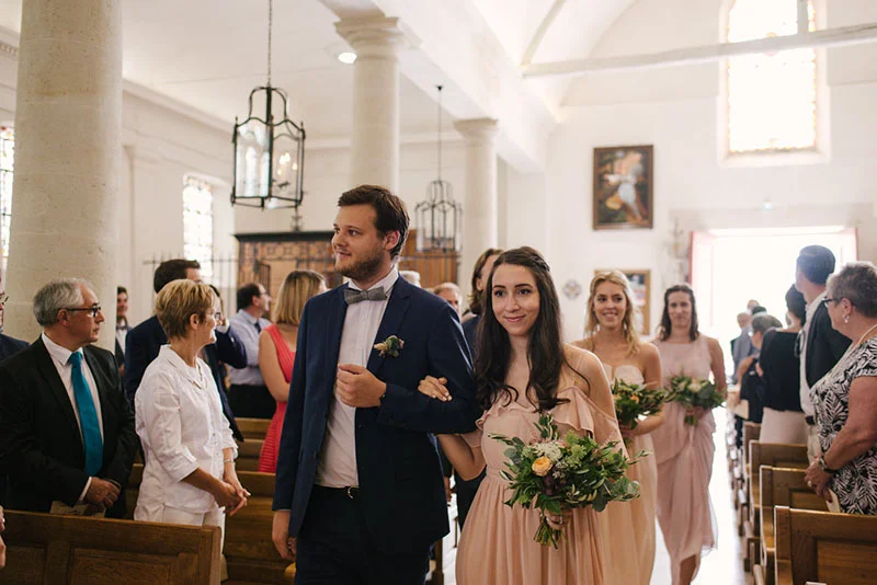 Un couple élégant avance dans l'allée d'une église lumineuse, entouré de leurs invités lors d'un mariage.