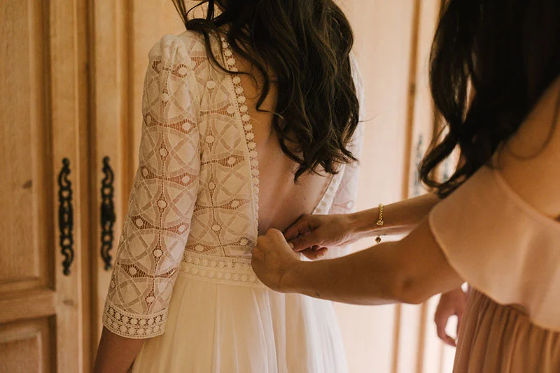 Une femme aide la mariée à fermer sa robe en dentelle blanche au dos dans une ambiance douce et lumineuse.