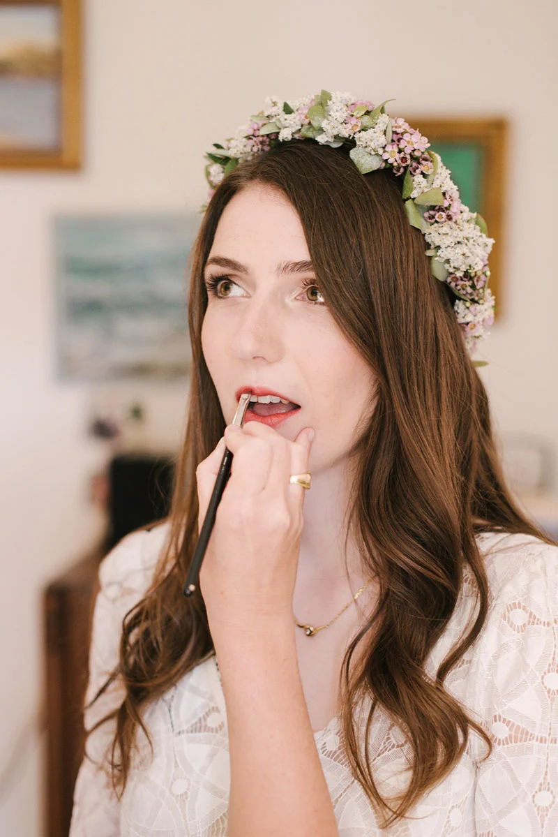 Une mariée aux cheveux longs portant une couronne de fleurs et une robe en dentelle blanche se maquille les lèvres.