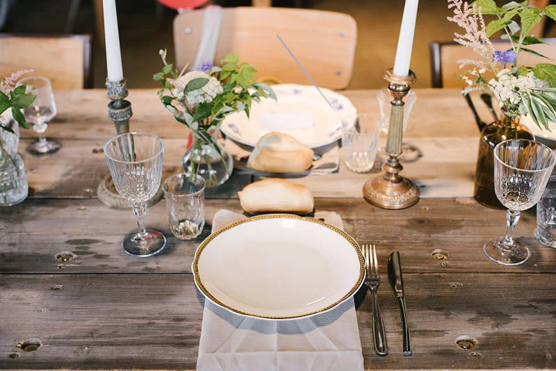 Table rustique en bois dressée avec vaisselle blanche, bougies et fleurs champêtres pour un mariage bohème.