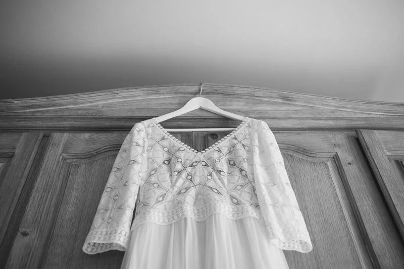 Robe de mariée blanche en dentelle suspendue devant un meuble en bois, photographie en noir et blanc.