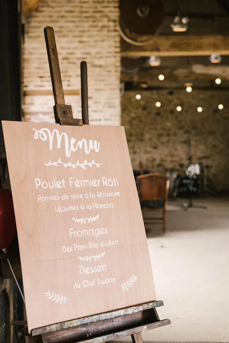 Menu de mariage écrit à la craie blanche sur un panneau en bois posé sur un chevalet dans une salle rustique.