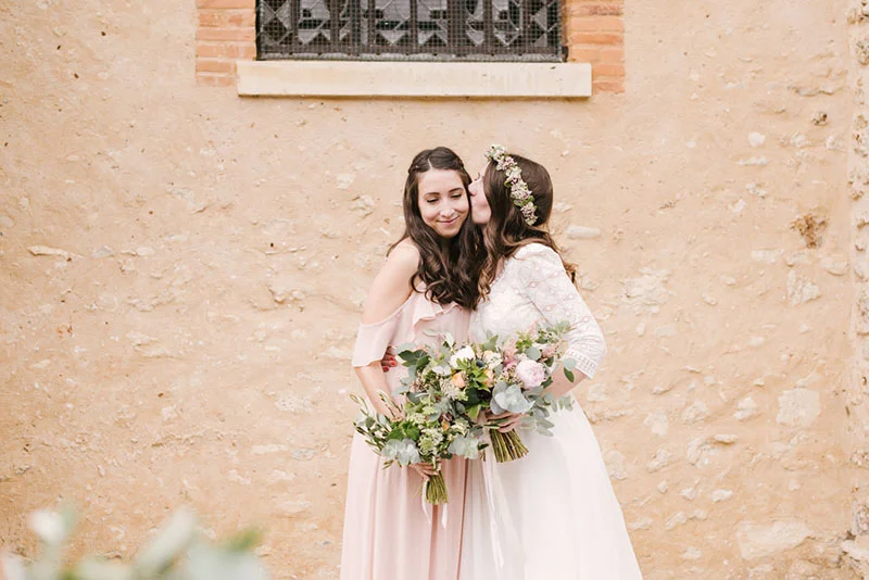 Deux femmes en robes rose pâle s'embrassent tendrement, tenant des bouquets de fleurs devant un mur en pierre.