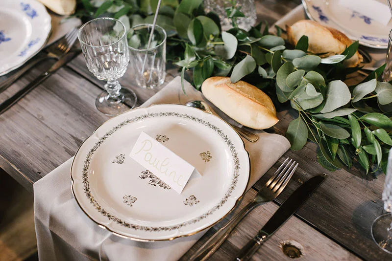 Table de mariage rustique avec assiette blanche, marque-place au nom de Pauline et décoration végétale en eucalyptus.