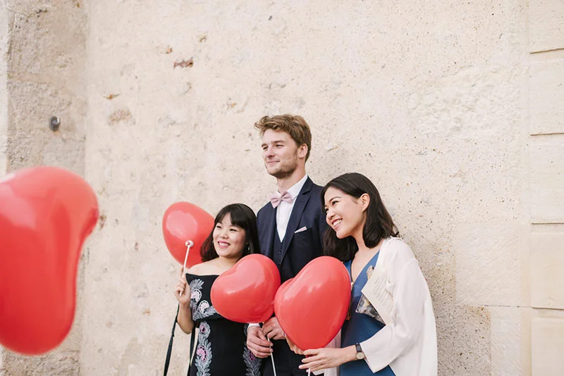 Un couple et une amie sourient avec des ballons rouges devant un mur en pierre claire dans une ambiance festive.