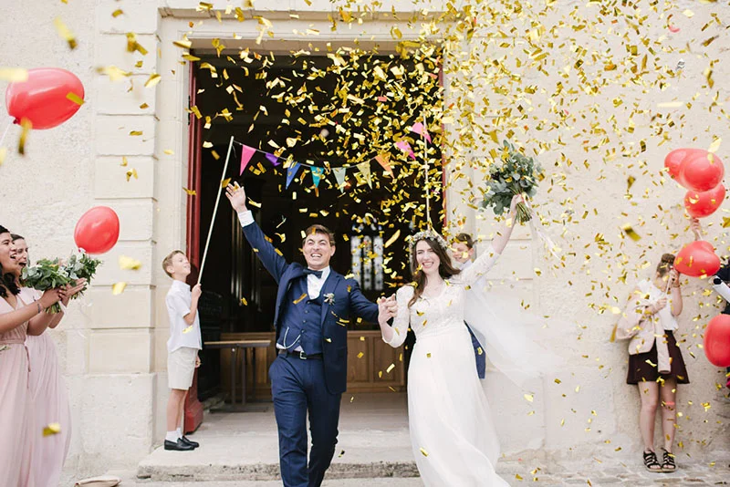 Mélanie et Jonathan sortent rayonnants de l'église sous une pluie de confettis dorés et des ballons rouges.