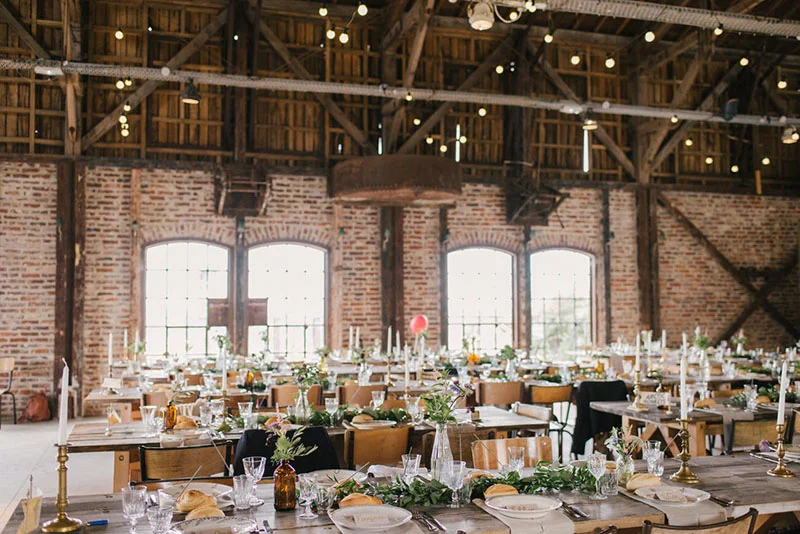 Réception de mariage champêtre dans une grange aux murs de briques avec tables en bois et décorations végétales.