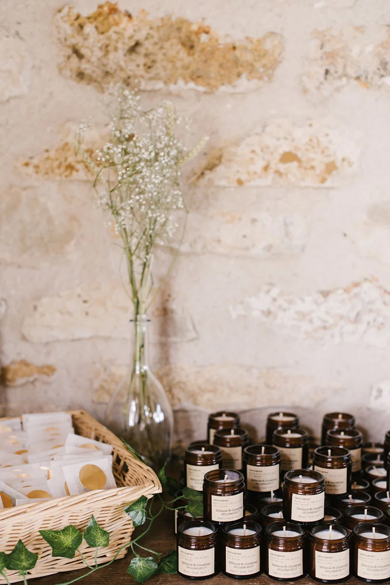 Cadeaux d'invités composés de bougies ambrées et sachets blancs devant un mur en pierre et un bouquet de gypsophile.