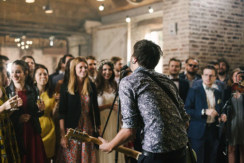 Un musicien à la guitare anime une réception de mariage chaleureuse devant des invités souriants dans un loft industriel.
