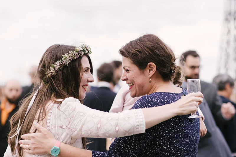 Deux femmes s'enlacent avec complicité lors d'un mariage, l'une portant une couronne de fleurs et une robe blanche.