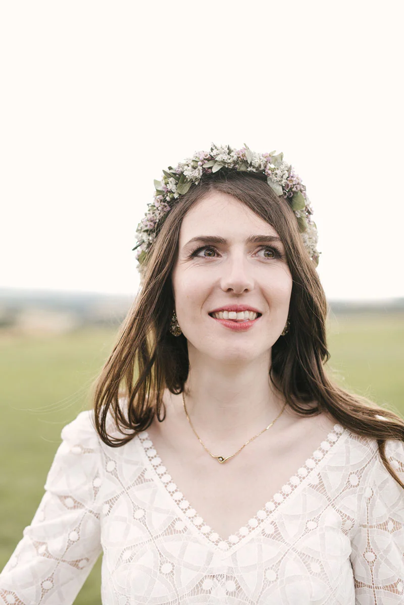 Portrait d'une mariée souriante portant une couronne de fleurs et une robe en dentelle blanche dans un champ naturel.