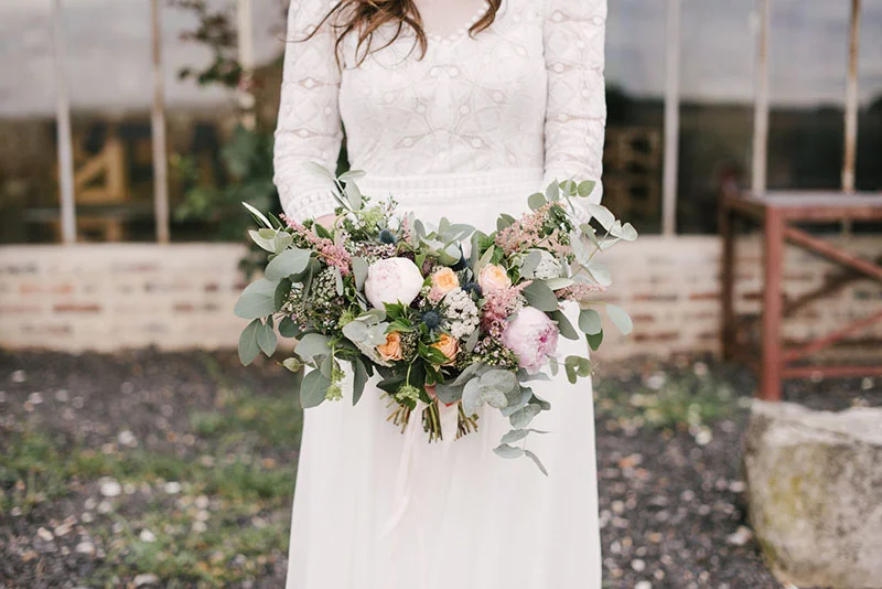Mariée en robe blanche dentelle tenant un bouquet champêtre aux tons pastel et feuillage d'eucalyptus.