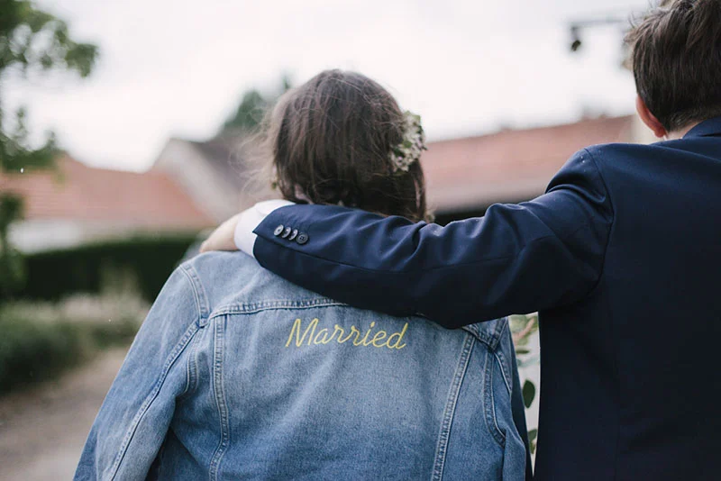 Un marié en costume bleu pose son bras sur l'épaule de sa femme portant une veste en jean brodée Married.
