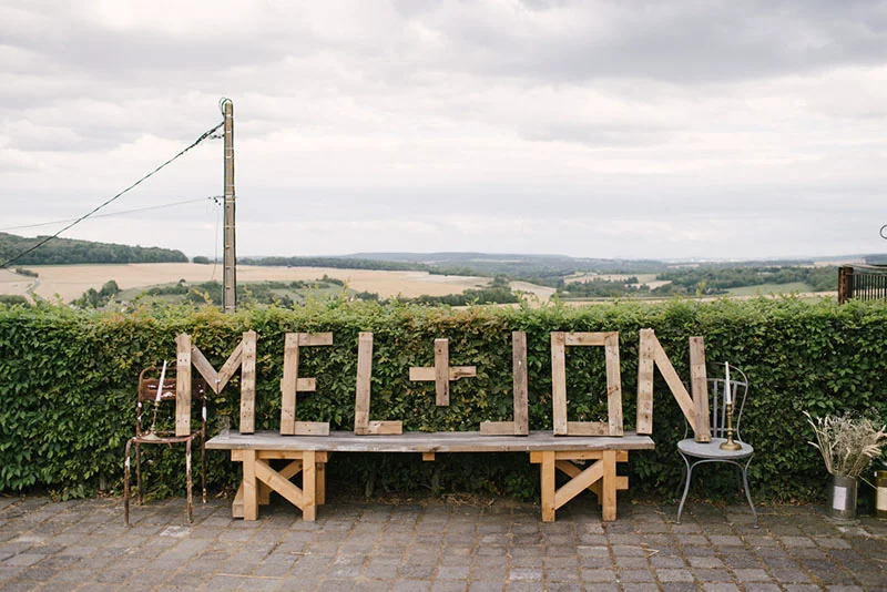 Lettres géantes en bois formant MEL + JON sur un banc rustique, face à un paysage champêtre sous un ciel nuageux.