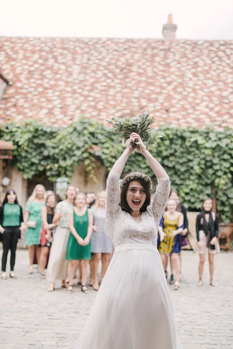 Une mariée joyeuse lance son bouquet devant ses invitées dans une cour rustique aux murs végétalisés.
