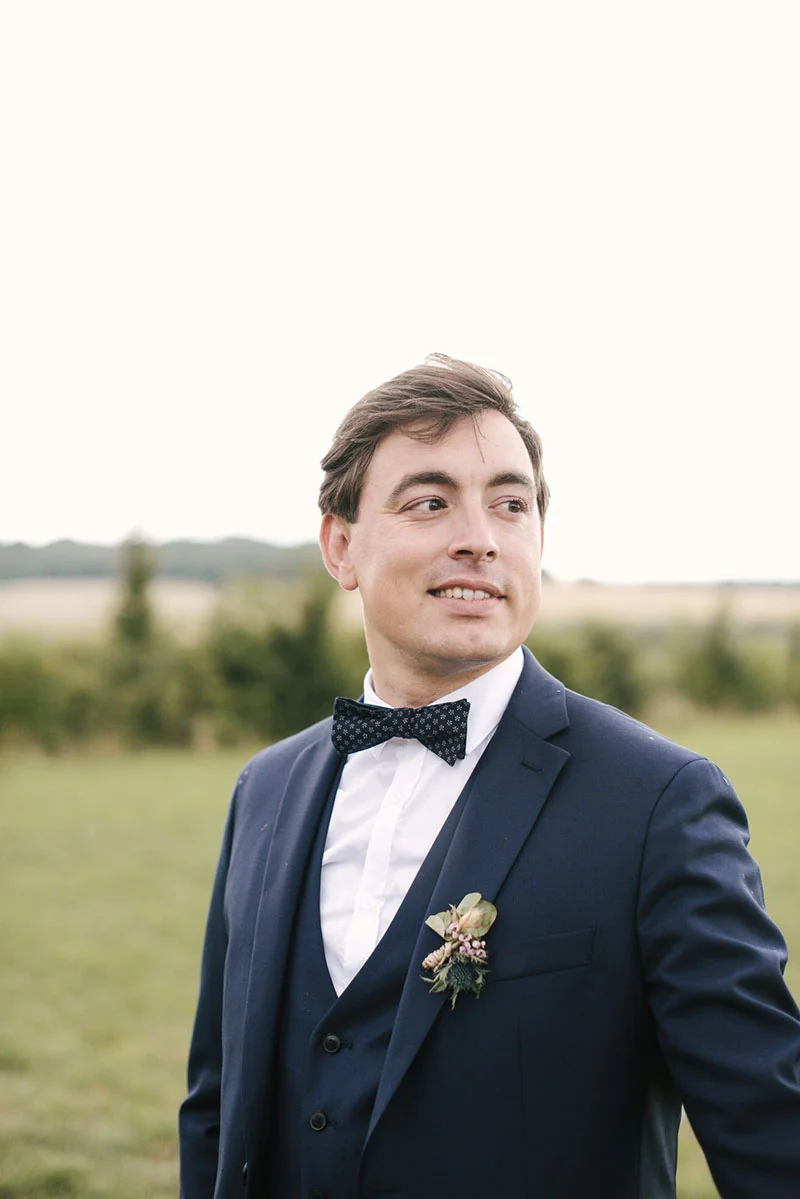 Portrait d'un marié souriant en costume bleu marine et nœud papillon, avec une boutonnière fleurie en extérieur.