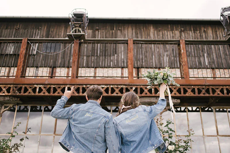 Jeunes mariés de dos en vestes en jean personnalisées devant un bâtiment industriel rustique.
