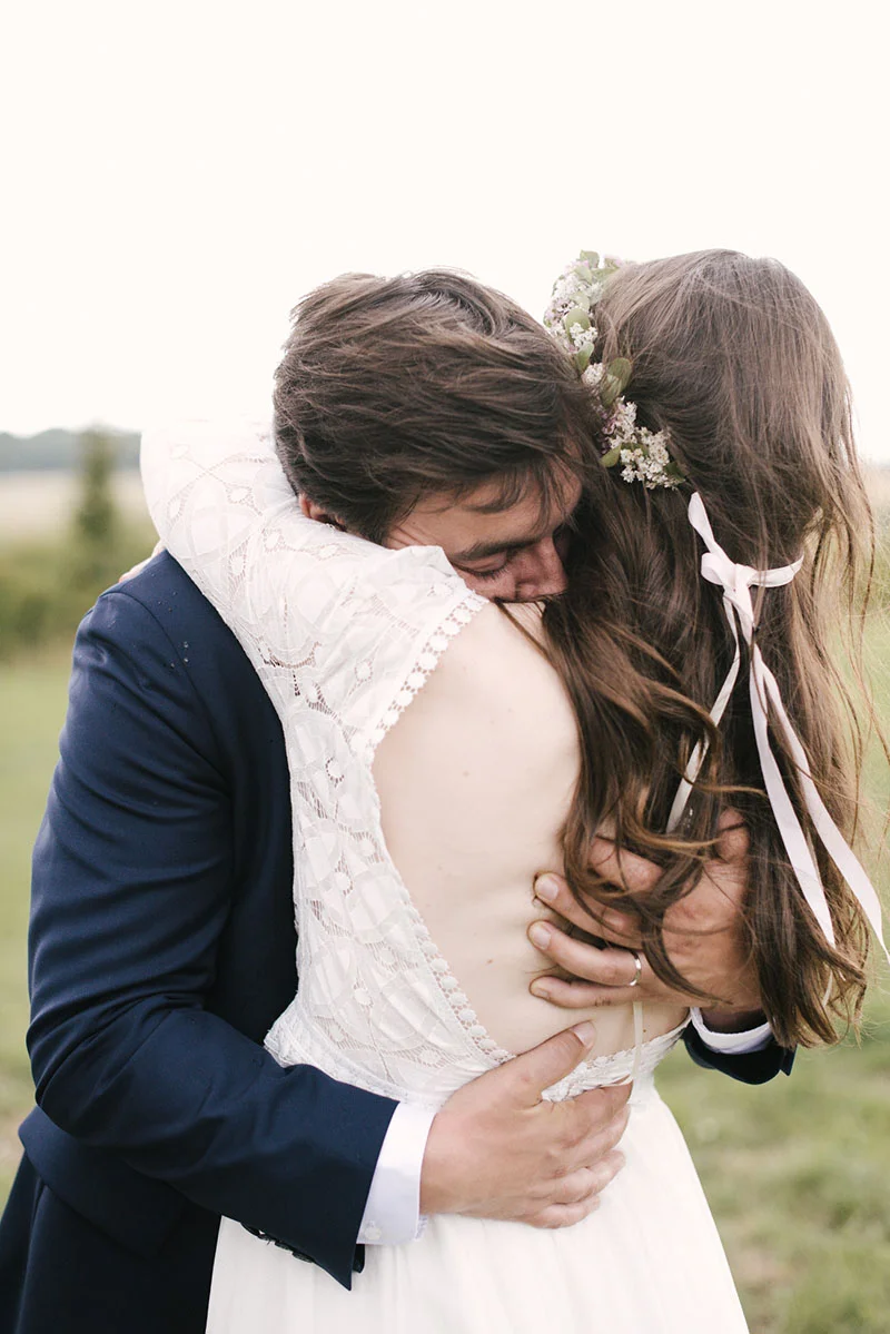 Jeune couple de mariés s'enlaçant tendrement en extérieur, elle en robe de dentelle blanche et lui en costume bleu marine.