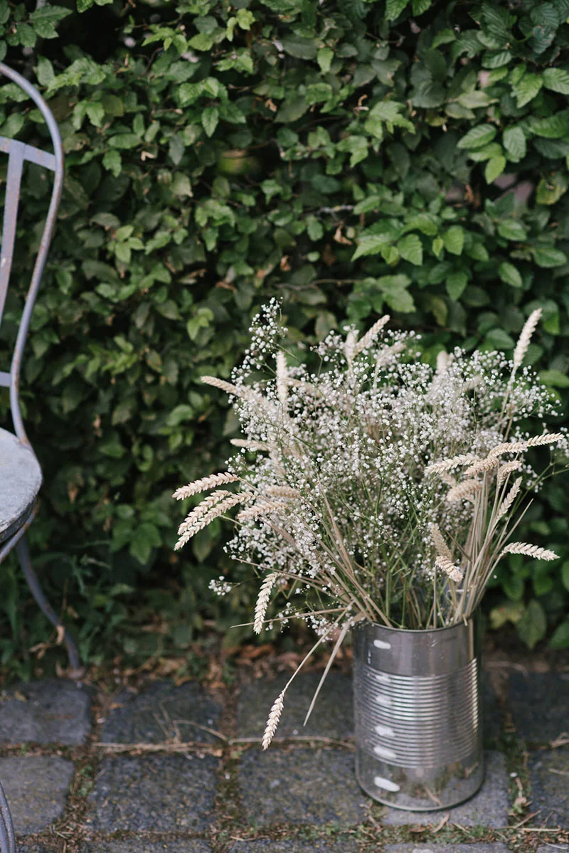 Bouquet de fleurs blanches et épis de blé dans une boîte de conserve argentée devant un mur de feuillage vert.