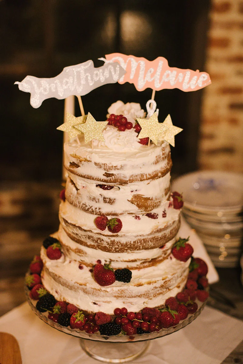 Gâteau de mariage naked cake à trois étages, décoré de baies rouges et d'étoiles dorées avec les prénoms des mariés.
