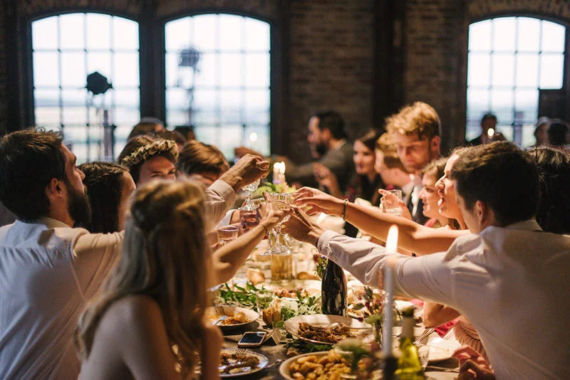 Invités trinquant joyeusement lors d'un dîner de mariage chaleureux dans un cadre industriel et lumineux.