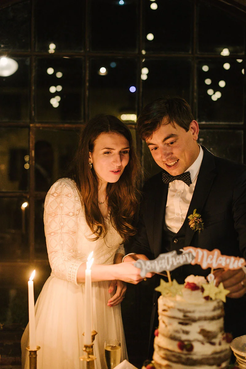 Jeunes mariés souriants posant un cake topper sur leur gâteau nu, éclairés à la bougie dans une ambiance chaleureuse.