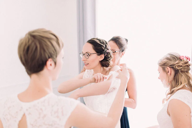 Une mariée à lunettes se fait aider à enfiler sa robe blanche par ses demoiselles d'honneur dans une ambiance lumineuse.