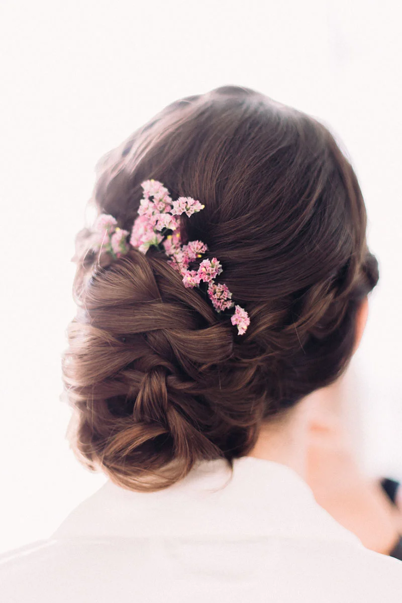Chignon bas romantic aux reflets châtains, orné de petites fleurs roses délicates.