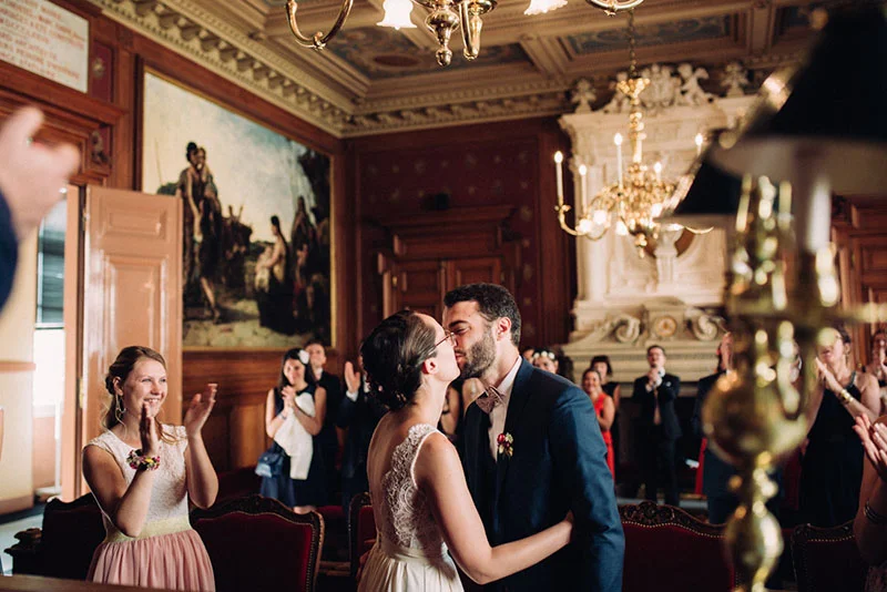 Clara et Julien s'embrassent lors de leur mariage dans un salon bourgeois chaleureux, entourés de leurs invités.