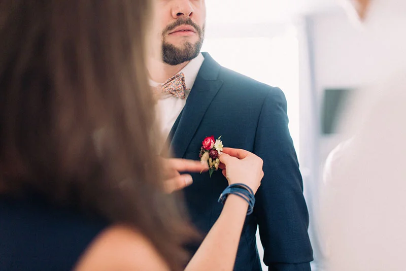 Une femme ajuste la boutonnière fleurie sur le costume bleu marine d'un homme portant un nœud papillon.