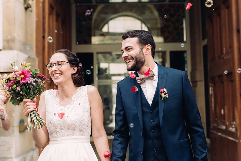 Jeunes mariés souriants sortant d'une église sous une pluie de bulles et de pétales, tenant un bouquet coloré.