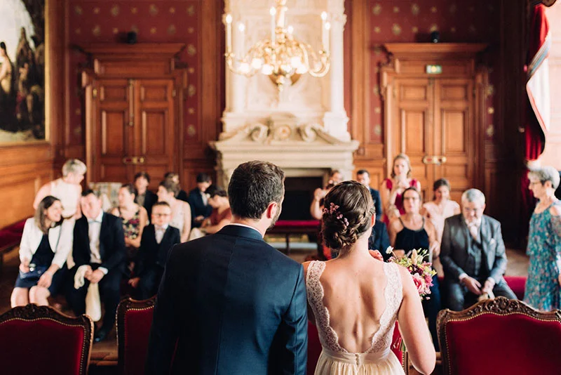 Mariés de dos face à leurs invités dans un salon bourgeois aux tons chauds et boiseries classiques.