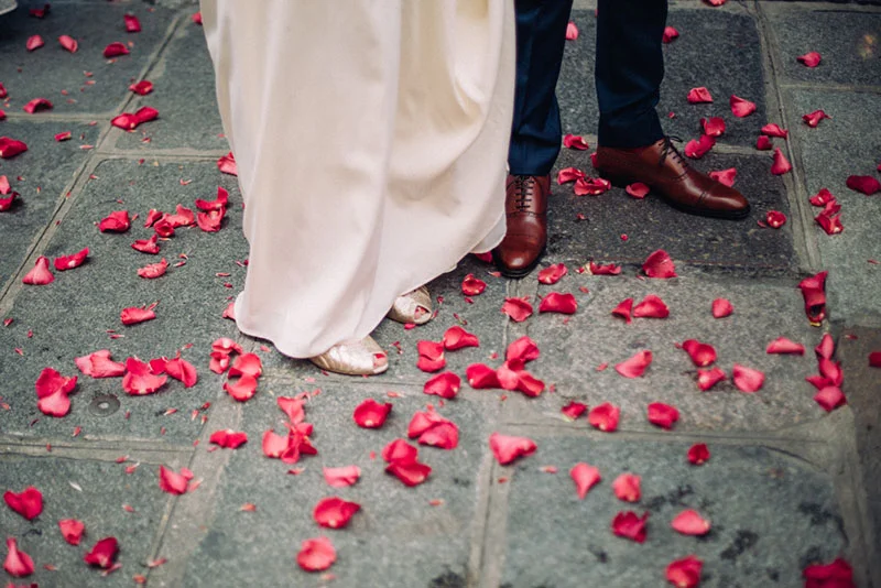 Gros plan sur les pieds d'un couple de mariés entourés de pétales de roses rouges sur un sol en pierre grise.