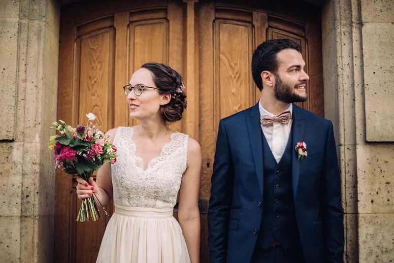 Jeunes mariés souriants devant une porte en bois, elle en robe blanche avec bouquet et lui en costume bleu marine.
