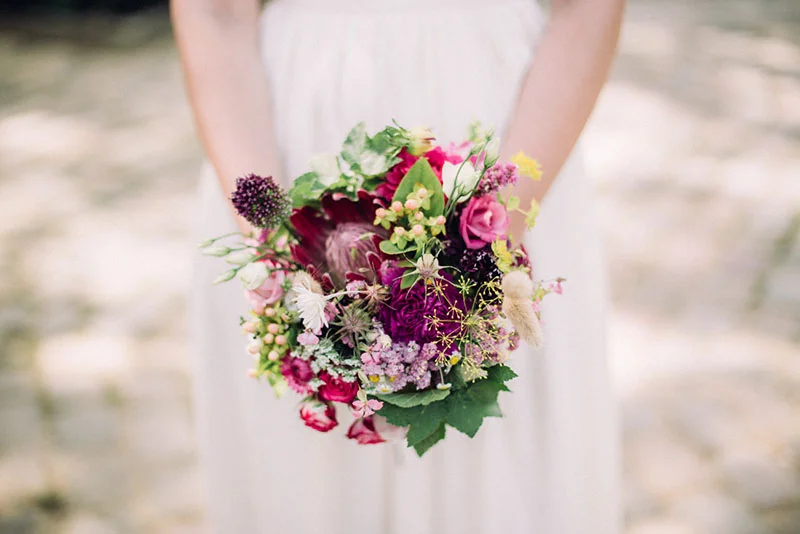 Une mariée en robe blanche tenant un bouquet champêtre aux nuances de rose, pourpre et vert.