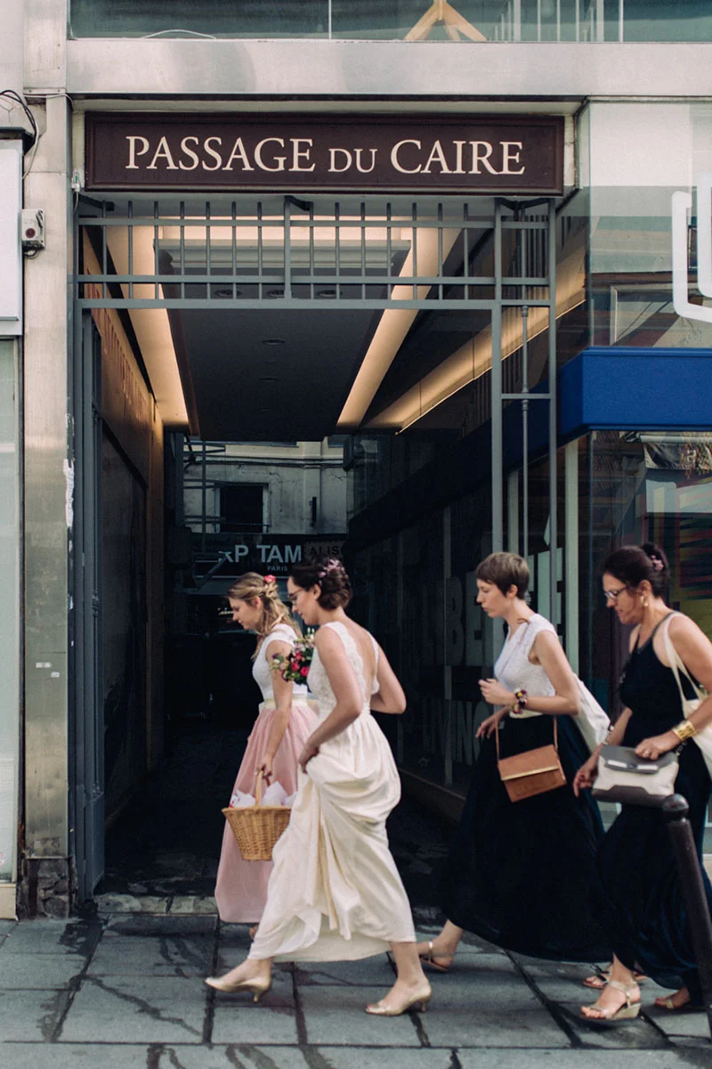 Trois femmes en robes élégantes, dont une mariée, marchent sous l'enseigne du Passage du Caire à Paris.