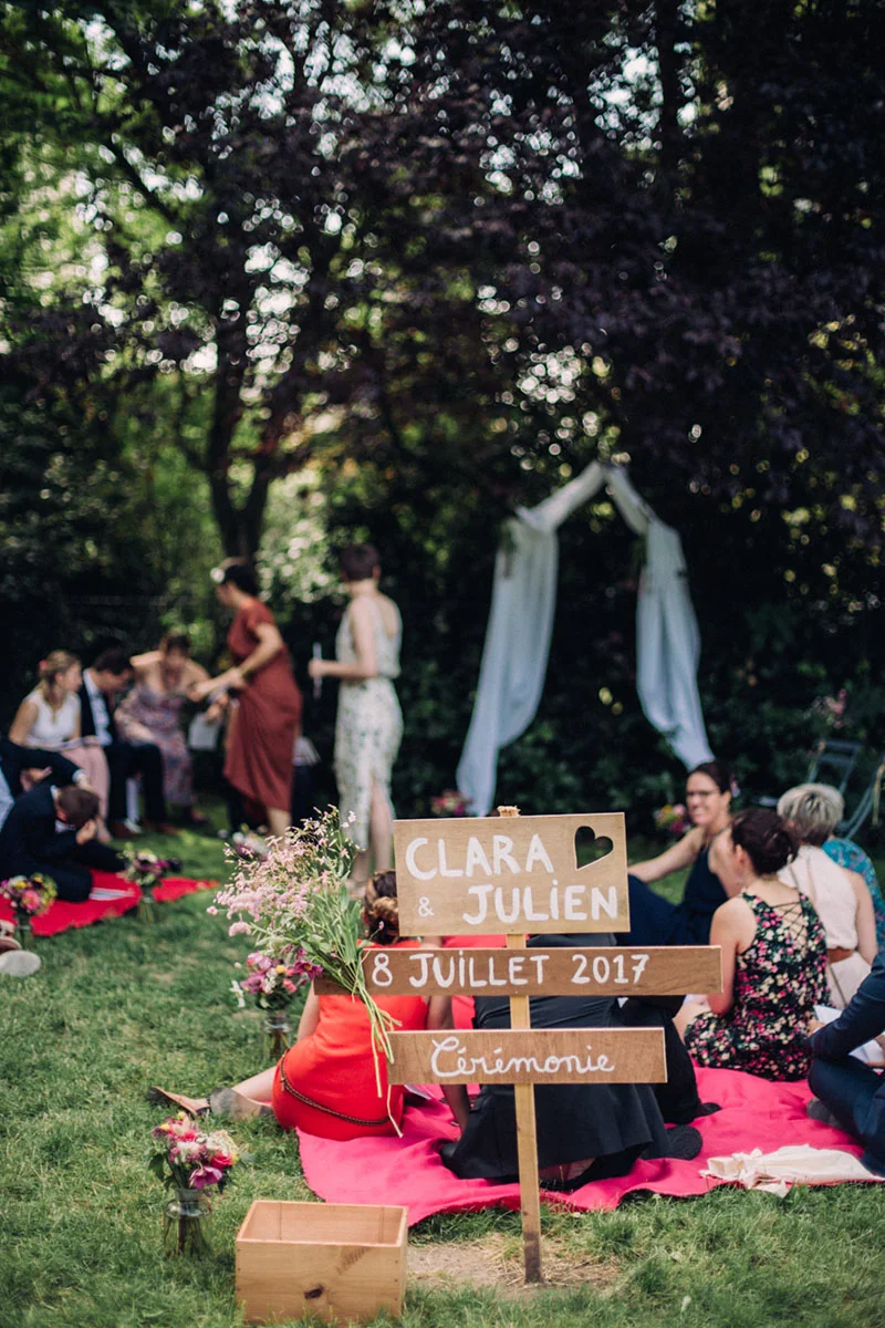 Panneau en bois pour le mariage de Clara et Julien dans un jardin verdoyant avec des invités sur des tapis rouges.