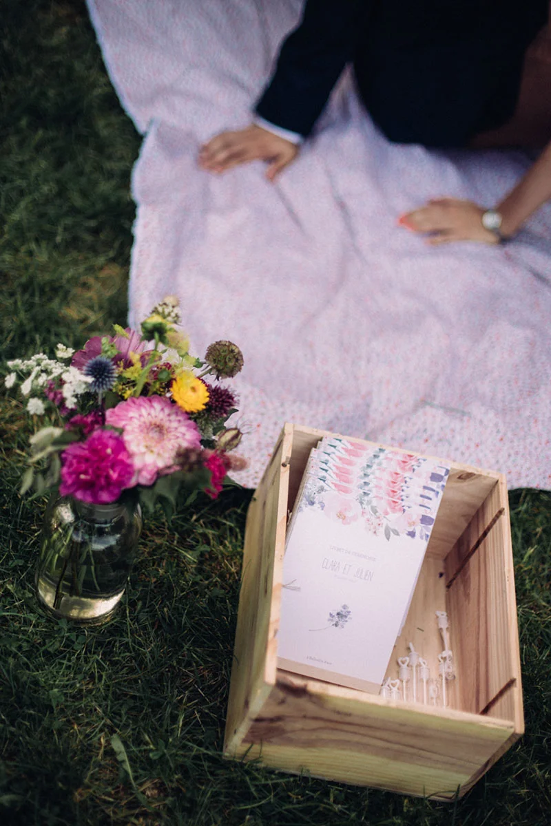 Bouquet de fleurs colorées et boîte en bois avec faire-part posés sur l'herbe à côté d'un couple sur un plaid.