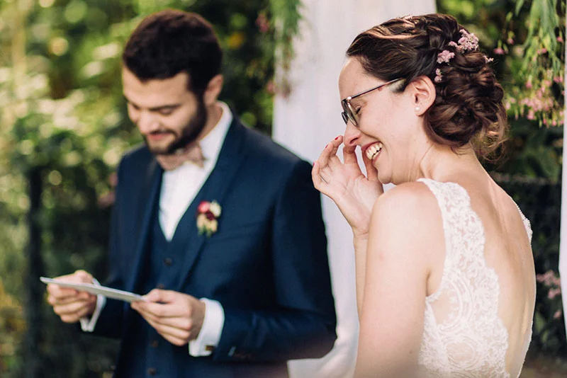 Un marié en costume bleu lit ses vœux à une mariée émue et souriante lors d'une cérémonie en plein air.