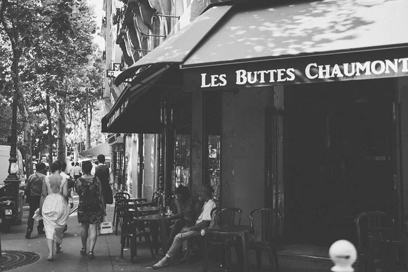 Photo noir et blanc d'une rue parisienne avec une terrasse de café et des passants, ambiance romantique et intemporelle.