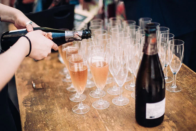 Une main verse du champagne rosé dans des flûtes alignées sur une table en bois rustique lors d'une réception.