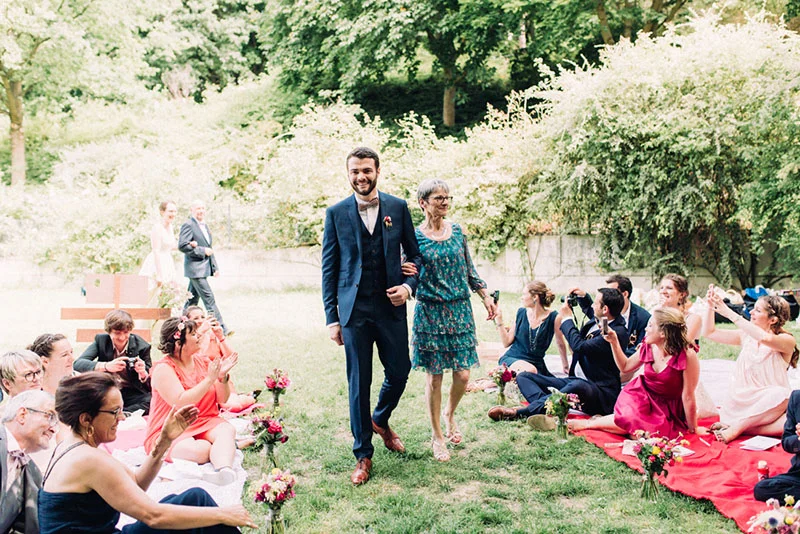 Julien, en costume bleu, marche au bras de sa mère vers des invités souriants lors d'une cérémonie en plein air.