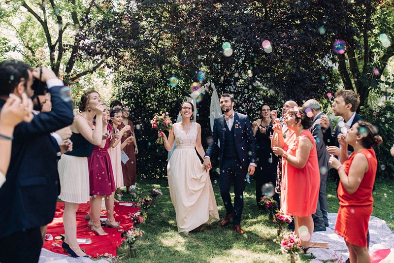 Jeunes mariés souriants marchant sous une pluie de bulles de savon entourés de leurs invités dans un jardin ensoleillé.