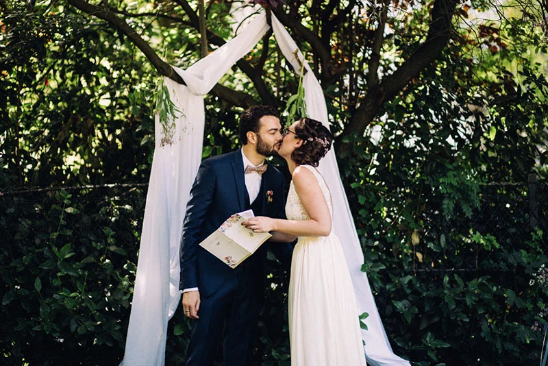 Un couple s'embrasse sous une arche de tissu blanc dans un jardin verdoyant lors de leur mariage.