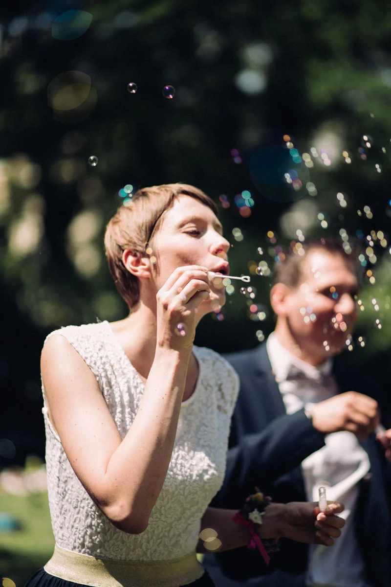 Une mariée en robe blanche souffle des bulles de savon à côté de son époux, dans une ambiance festive et lumineuse.