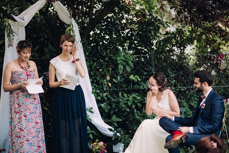 Une mariée émue et son époux écoutent deux amies prononcer des discours sous une arche blanche en plein air.