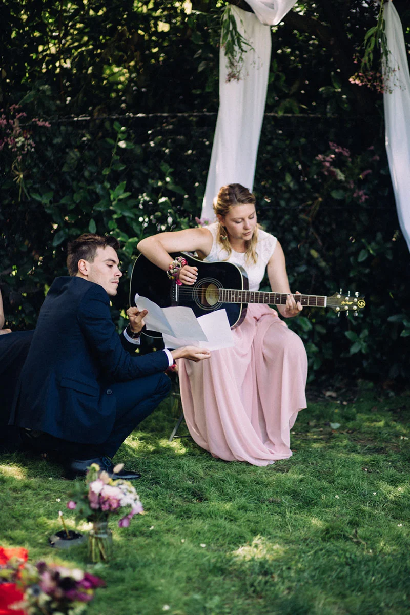 Une femme en robe rose joue de la guitare tandis qu'un homme en costume bleu lit un texte dans un jardin verdoyant.
