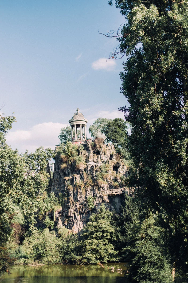 Un temple classique perché sur un rocher escarpé et verdoyant, entouré d'arbres sous un ciel bleu clair.
